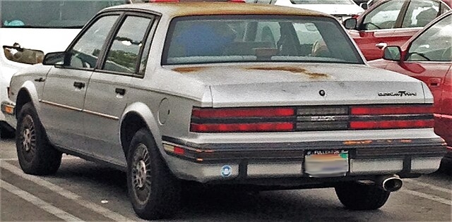 An old rusted Buick in a parking lot. The license plate is blurry.