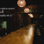 A view down a very long wood-grain bar with bar supplies on the left and several bar stools on the right. The bar is dimly lit by several overhead globe lamps. Text on the image: "I got an extra room in the back. Don't usually let it."